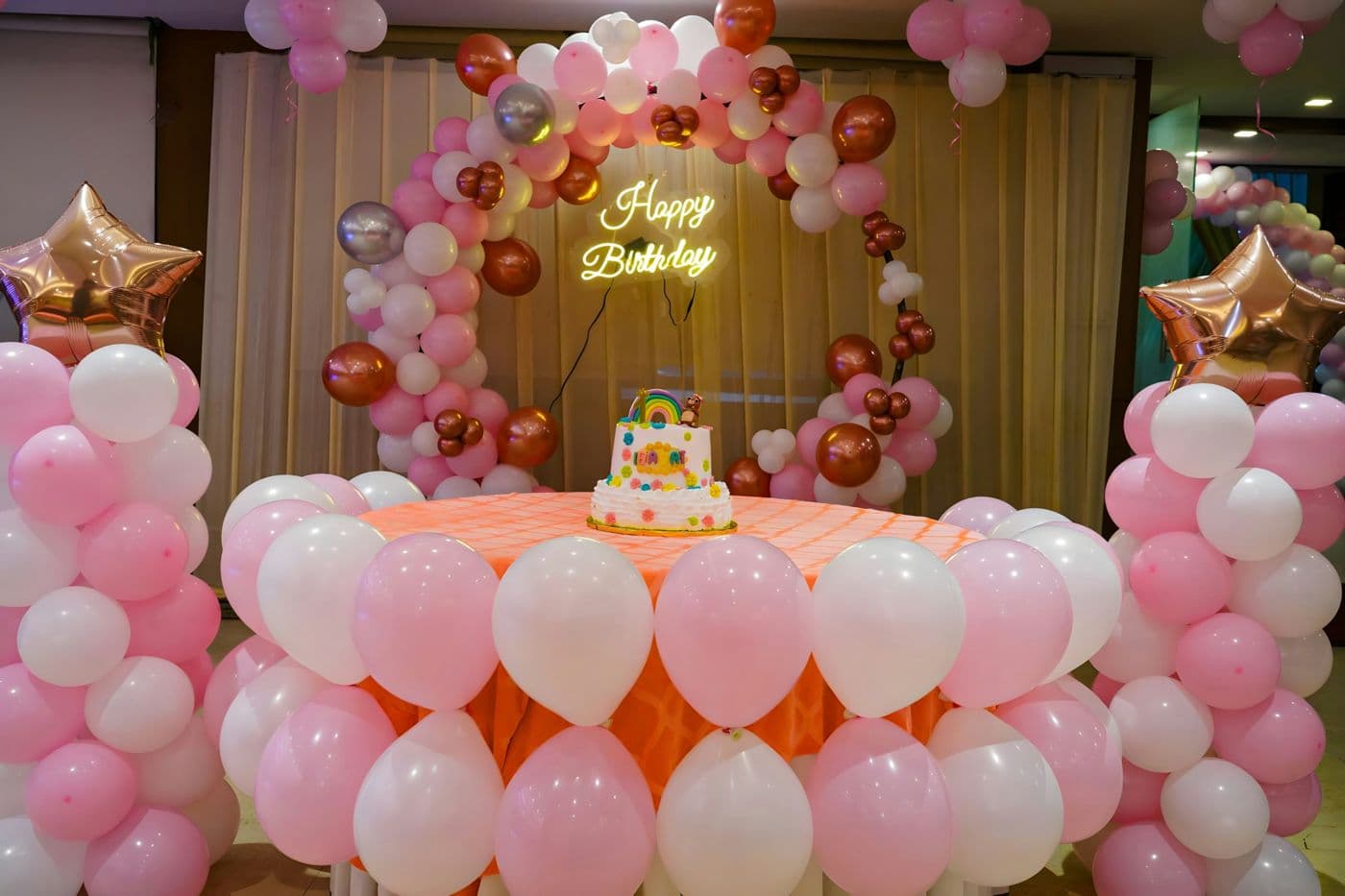 Child's birthday table with a cake, pink and white balloons and a Happy Birthday neon sign
