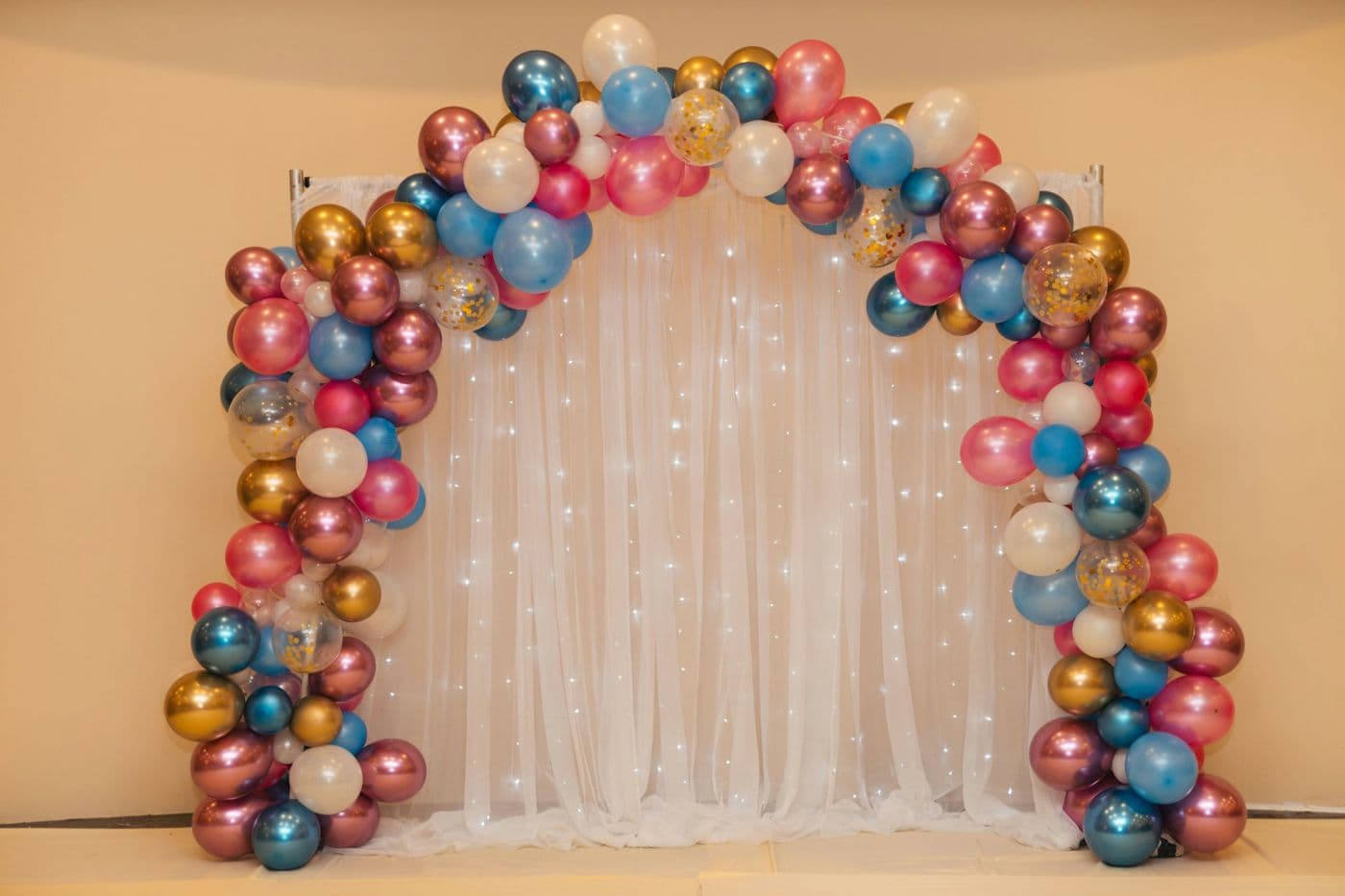 Arche de ballons roses, bleus et dorés devant un rideau lumineux