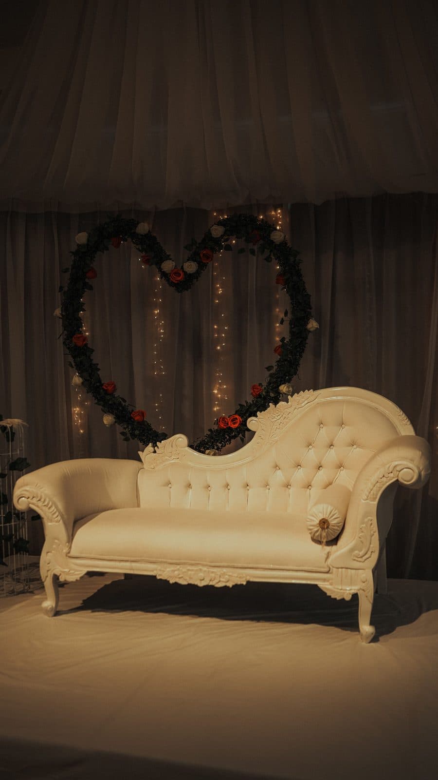 White chaise longue in front of a floral heart and fairy-light curtain